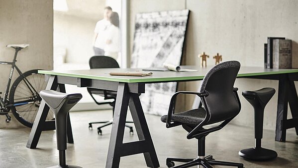 Besprechung in einem modernen Büro mit Stehpult und Sitzbock, Mitarbeitende im Austausch.
