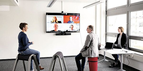 Videokonferenz mit mehreren Teilnehmern: zwei Teilnehmer sitzen auf modernen Sitzhockern vor einem Bildschirm.