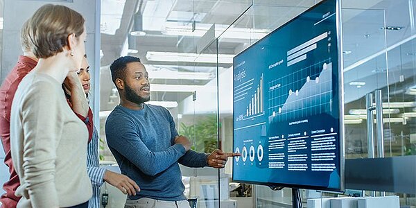 Digitale Präsentation, Team bespricht Datenanalyse vor einem Bildschirm mit Diagrammen und Statistiken.