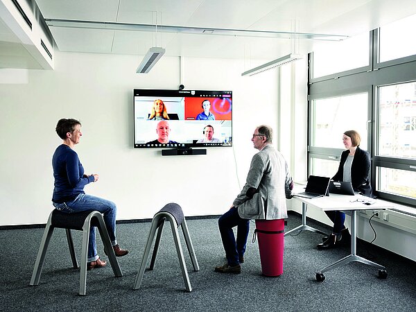 Videokonferenz mit mehreren Teilnehmern: zwei Teilnehmer sitzen auf modernen Sitzhockern vor einem Bildschirm.